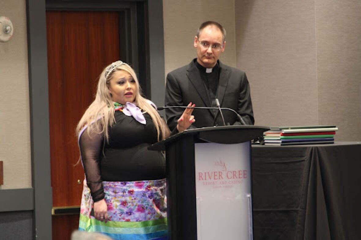 A man wearing glasses and a Roman collar stands at a podium alongside a woman with long hair, a black shirt and a colourful skirt. 